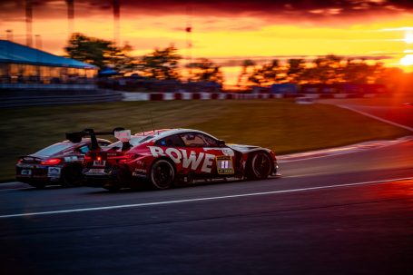 Rennwagen mit Sponsorenlogo fährt bei Sonnenuntergang auf einer Rennstrecke.