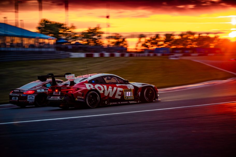Rennwagen mit Sponsorenlogo fährt bei Sonnenuntergang auf einer Rennstrecke.